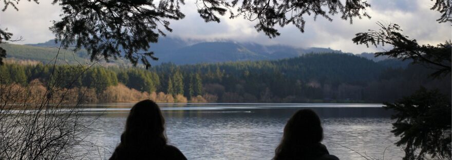 Photograph of the silhouette of the backs of two ladies sitting on a bench loooking out over a lake, trees and hills in the background