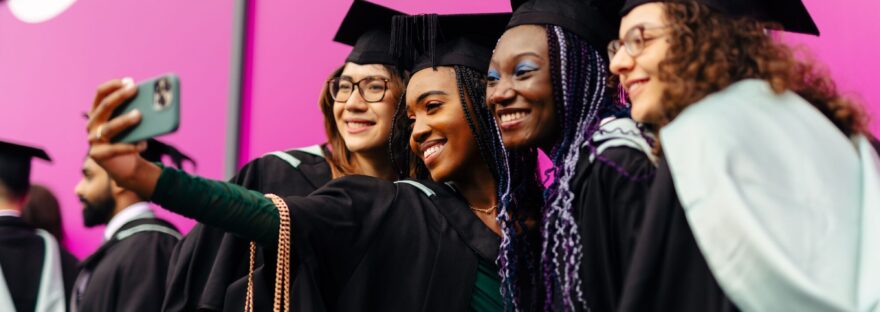 Photograph of four graduates in gowns and hoods taking a selfie
