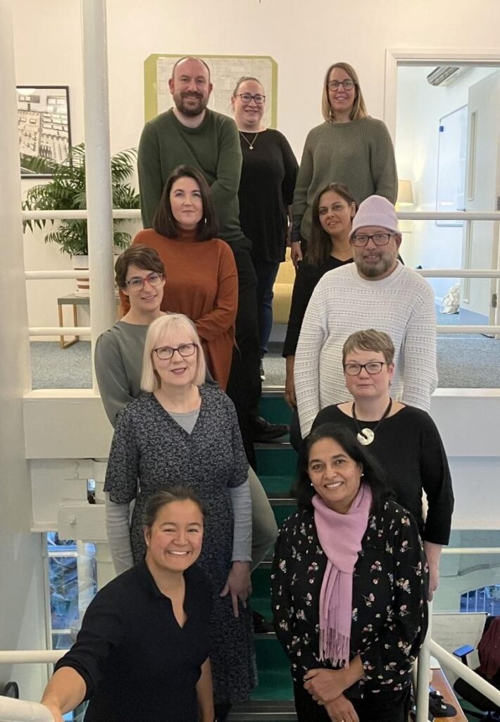 The image shows a diverse group of people - some members of the In It Together working group - standing on a staircase. They are mostly smiling.
